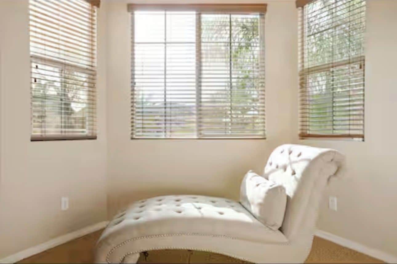 Una habitación de esquina con una butaca tapizada de blanco, una ventana con persianas y suelo alfombrado.