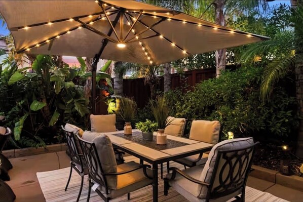 Une terrasse extérieure avec une table et des chaises, entourée d’une végétation luxuriante et agrémentée d’un grand parasol décoré de guirlandes lumineuses.