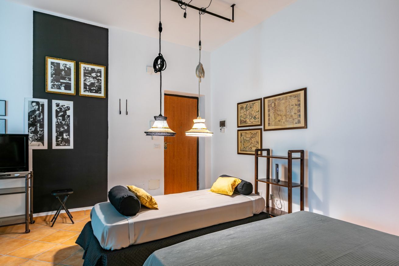 Habitación pequeña con cama, escritorio y televisor. En las paredes hay cuadros enmarcados y una puerta que conduce a otra habitación.