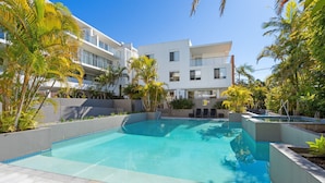 A modern pool area with a clear blue pool, surrounded by palm trees and a multi-story building with balconies. - Riverside luxury apartment across two levels at Waterline. (Forster)