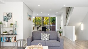 A modern living room with a grey sofa, a bookshelf, and a dining area visible through large windows. - Riverside luxury apartment across two levels at Waterline. (Forster)