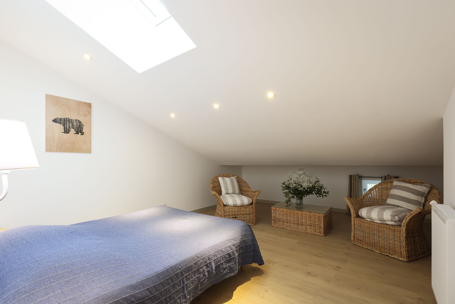 A bedroom with a bed, two wicker chairs, a small table with a vase, and a ceiling light.