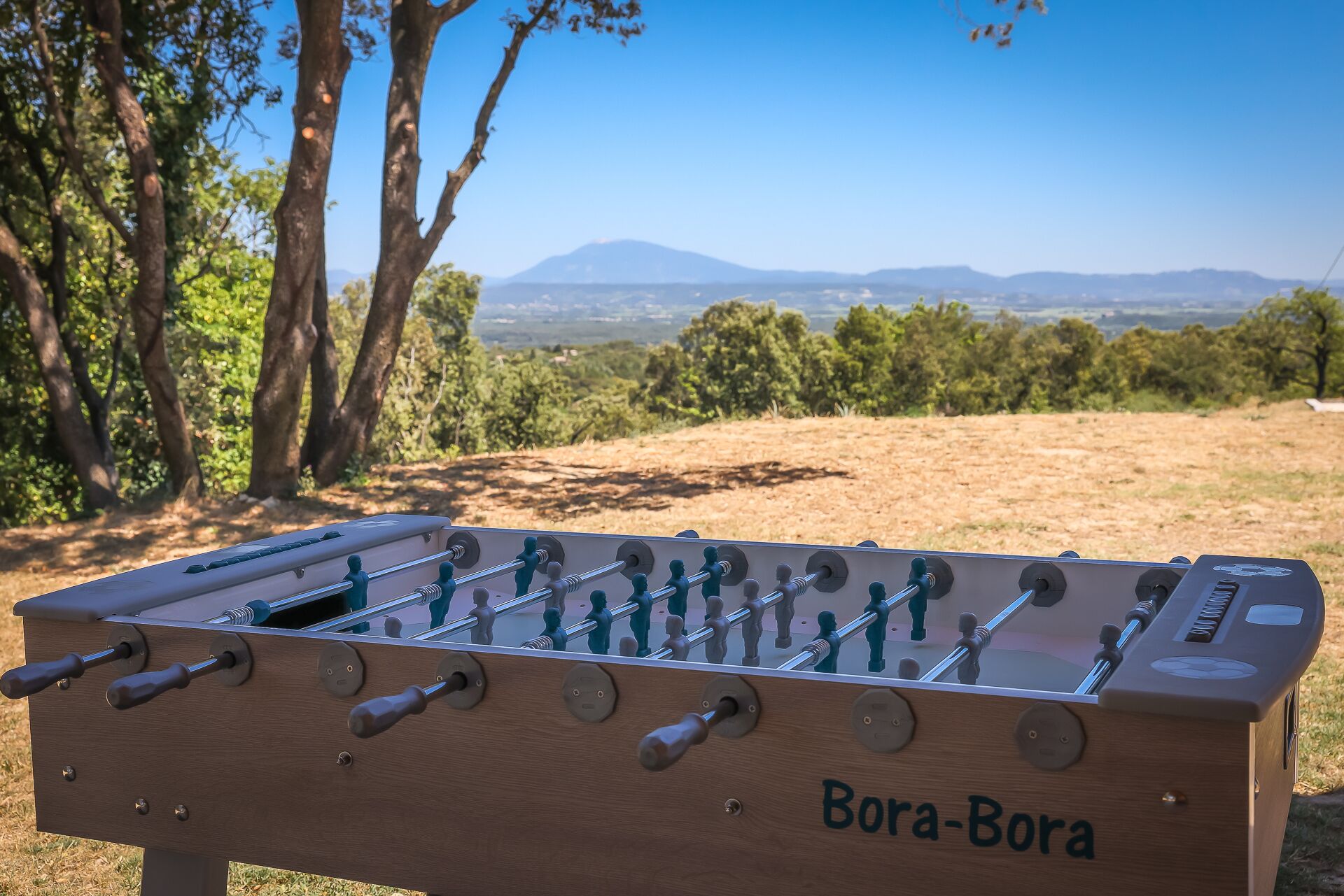 A wooden foosball table with the name "Bora-Bora" on it, set against a backdrop of trees and a distant mountain range.