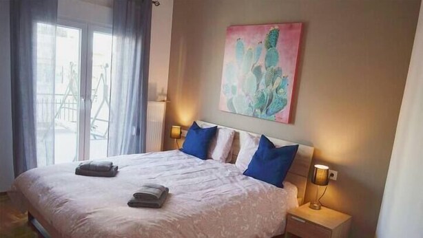 A bedroom with a large bed, two bedside lamps, a painting on the wall, and a view through the window. - GTD33702 - Feel the Acropolis Apt (Athens)