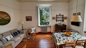 Uma sala de estar aconchegante com um sofá, uma mesa de jantar com toalha florida, uma cadeira de vime, uma mesa de centro de vidro e uma janela com vista para o verde.