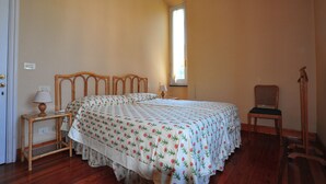 Quarto com cama, mesa de cabeceira com luminária, cadeira e janela.