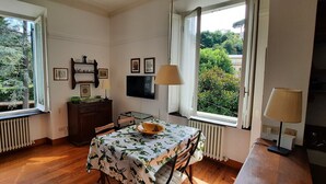 Quarto com mesa de jantar, cadeiras, televisão e janela com vista para vegetação.