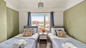 Ein Doppelzimmer mit zwei Betten, einem Fenster mit Vorhängen, einem Nachttisch mit Lampe und Blick auf Gebäude.