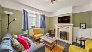 Ein Wohnzimmer mit einer Couch, Sesseln, einem Couchtisch, einem Kamin und einem Fernseher, der 'NETFLIX' anzeigt.