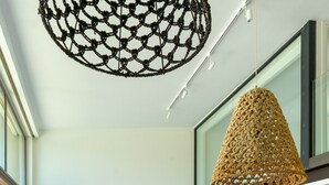 Uma sala de estar moderna com uma mesa de jantar redonda de madeira, sofá cinza com almofadas laranja e uma luminária pendente grande e trançada.