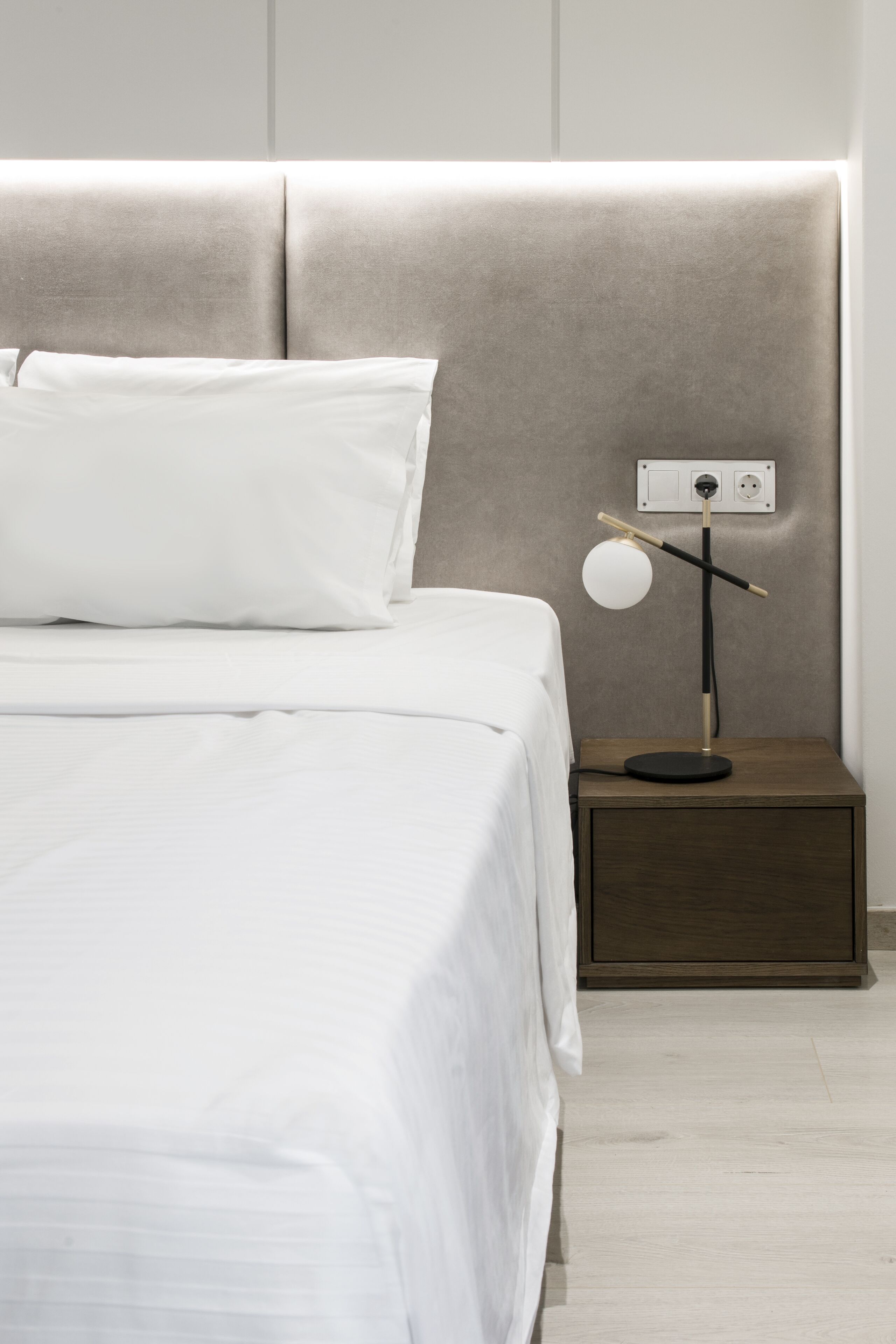 Un letto rifatto con lenzuola bianche, un comodino con una lampada e una presa di corrente a muro.