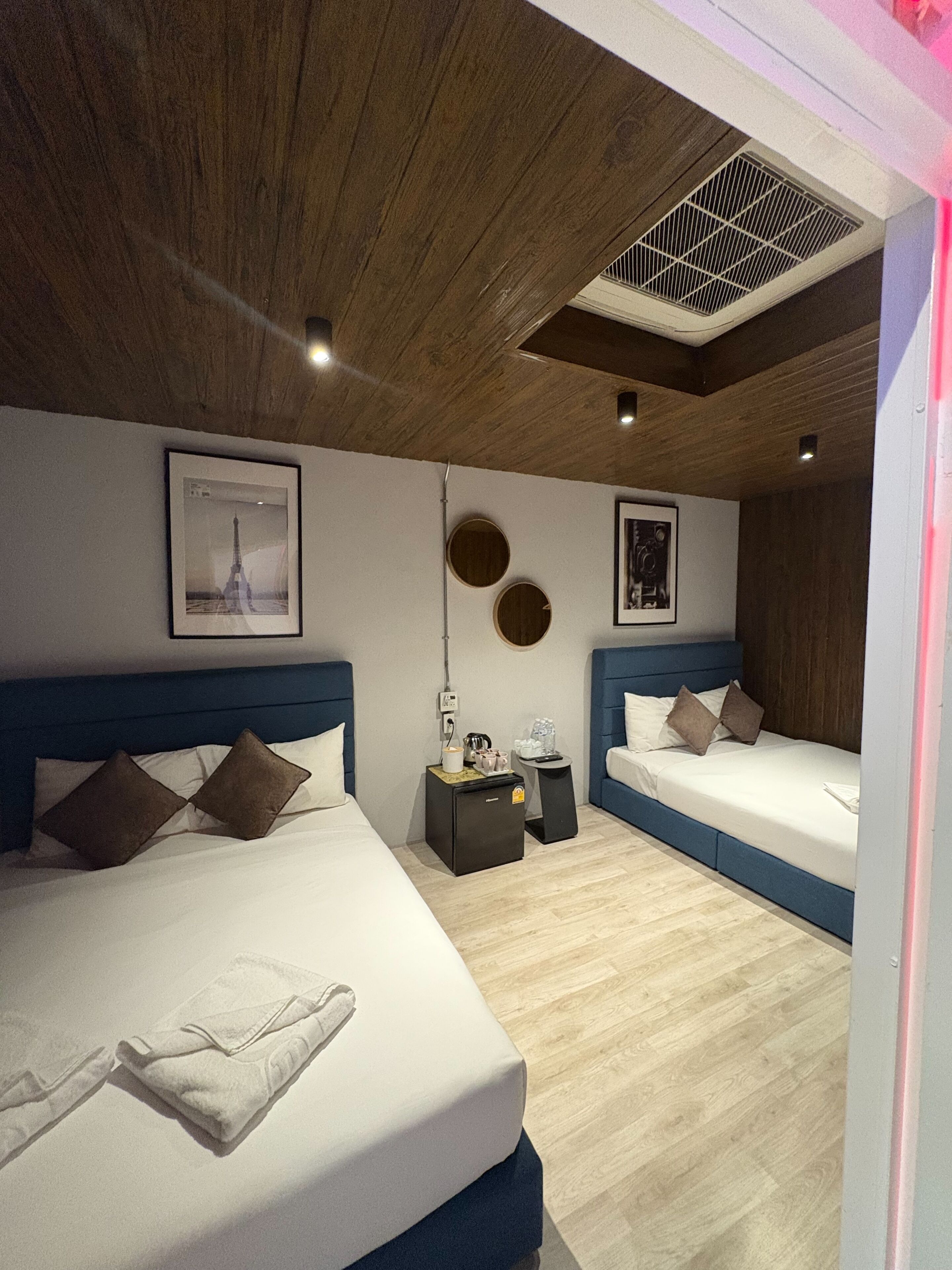 A modern hotel room with two beds, wooden ceiling, and a small kitchenette.