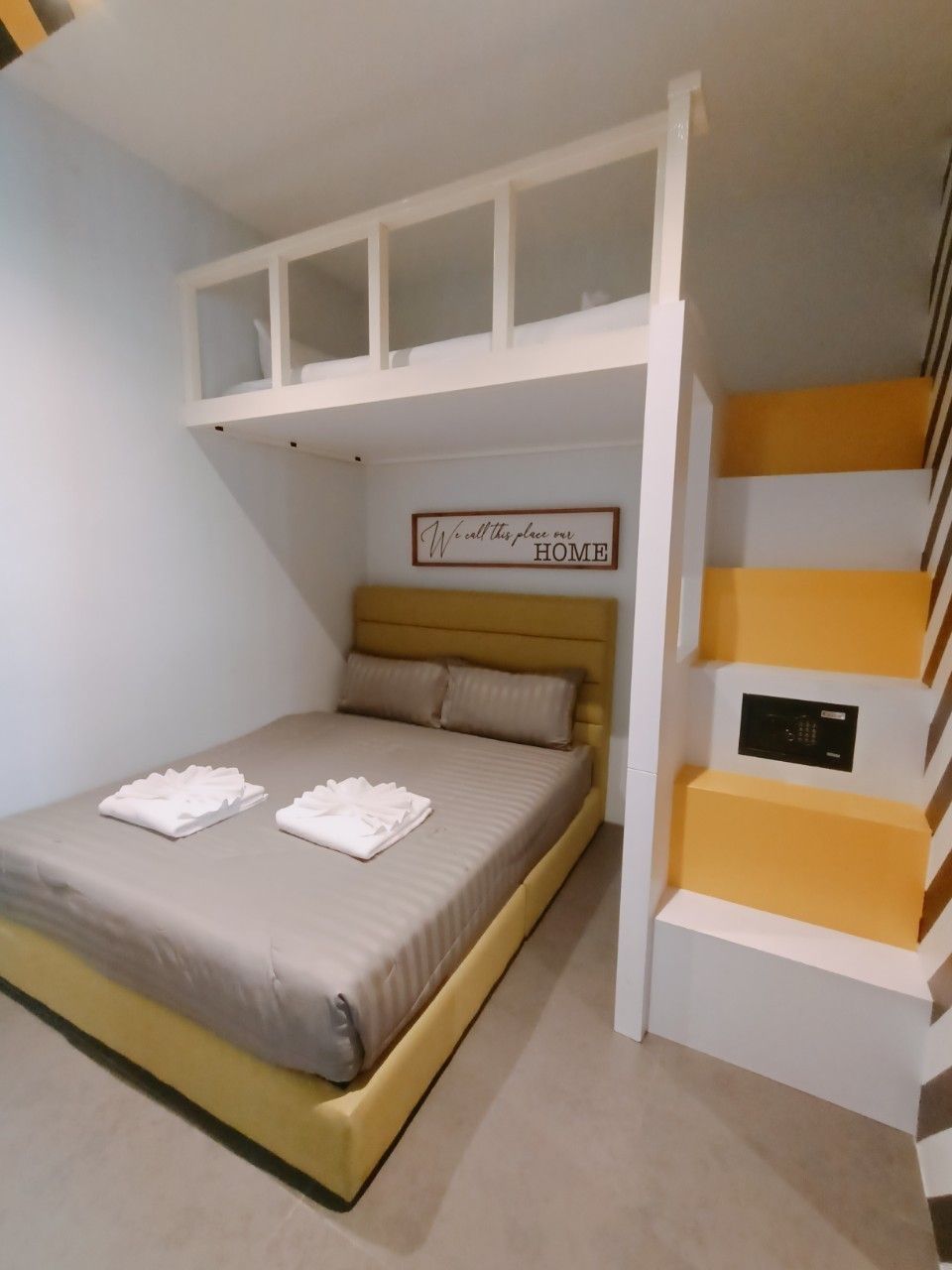 Une chambre avec un lit superposé, un lit avec deux oreillers, une table de chevet dotée d’un panneau de commande, et une pancarte au-dessus du lit sur laquelle on peut lire : « Nous appelons cet endroit notre FOYER. »
