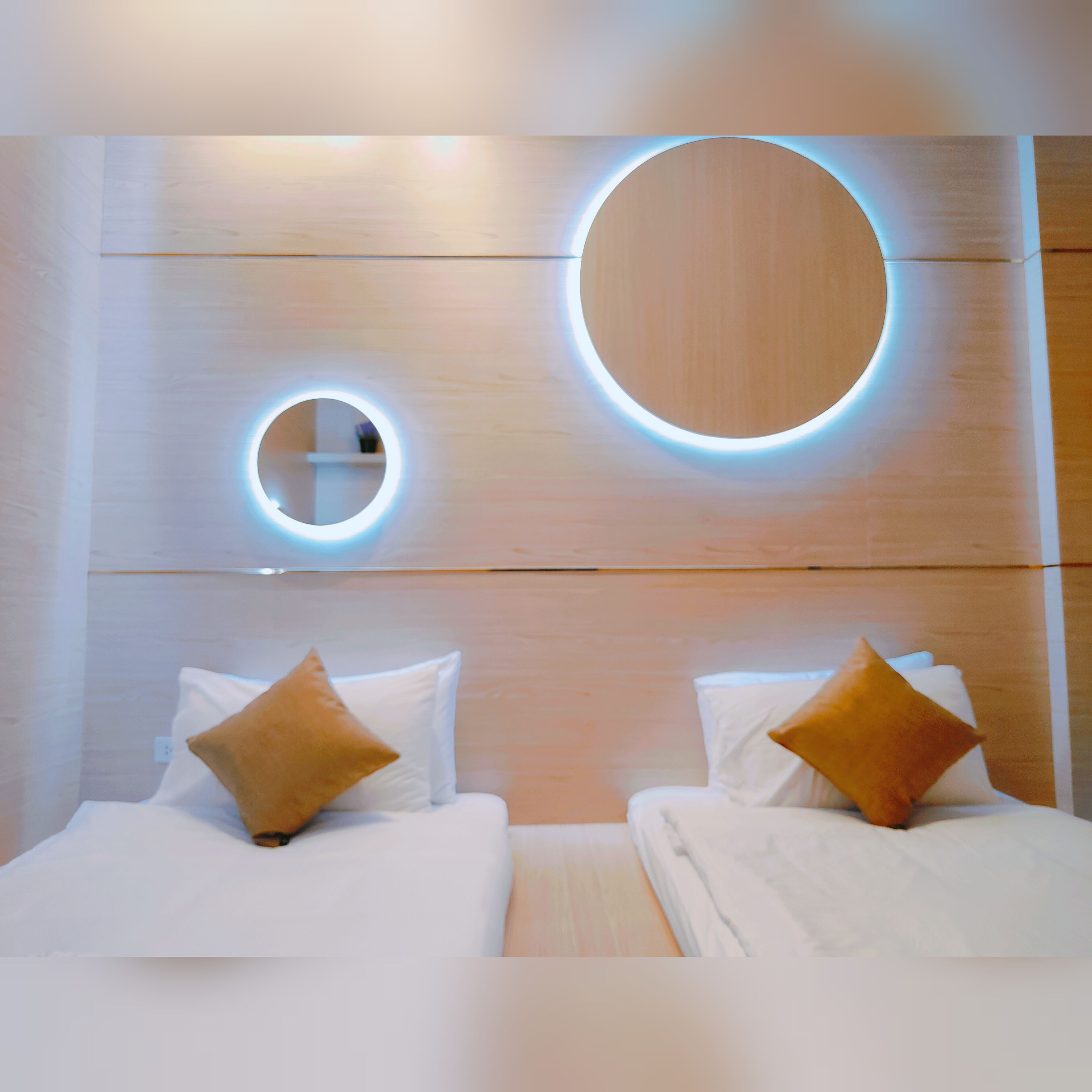 Una camera d'albergo moderna con due letti, ognuno con biancheria da letto bianca e un cuscino marrone. Sopra i letti, sono presenti lampade circolari a parete.