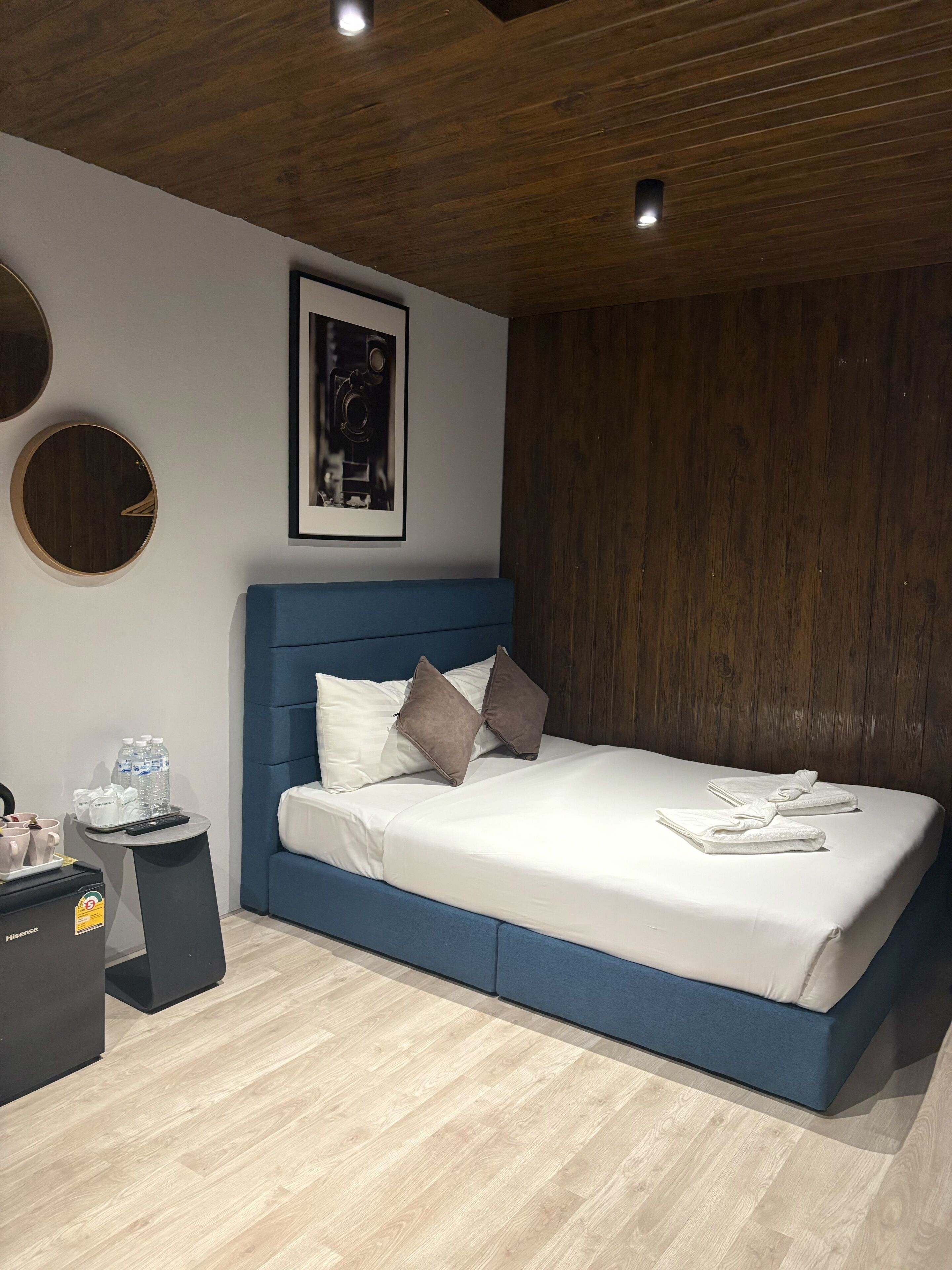 Quarto moderno com cama azul, roupa de cama branca e cabeceira de madeira. Há um quadro na parede e uma pequena mesa com um controle remoto e garrafas de água.
