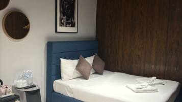 Quarto moderno com cama azul, roupa de cama branca e cabeceira de madeira. Há um quadro na parede e uma pequena mesa com um controle remoto e garrafas de água.