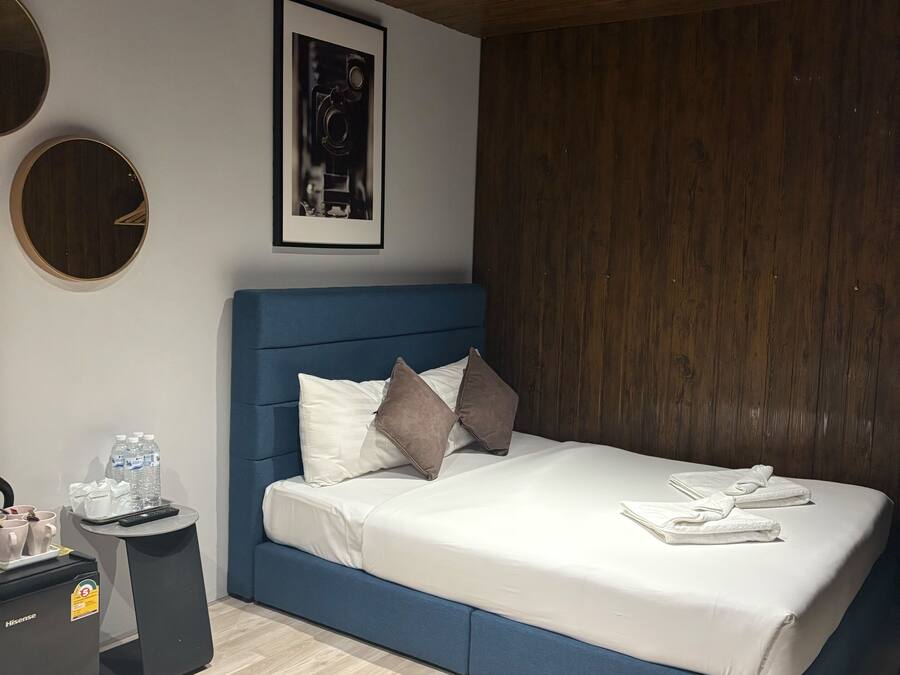 Quarto moderno com cama azul, roupa de cama branca e cabeceira de madeira. Há um quadro na parede e uma pequena mesa com um controle remoto e garrafas de água.