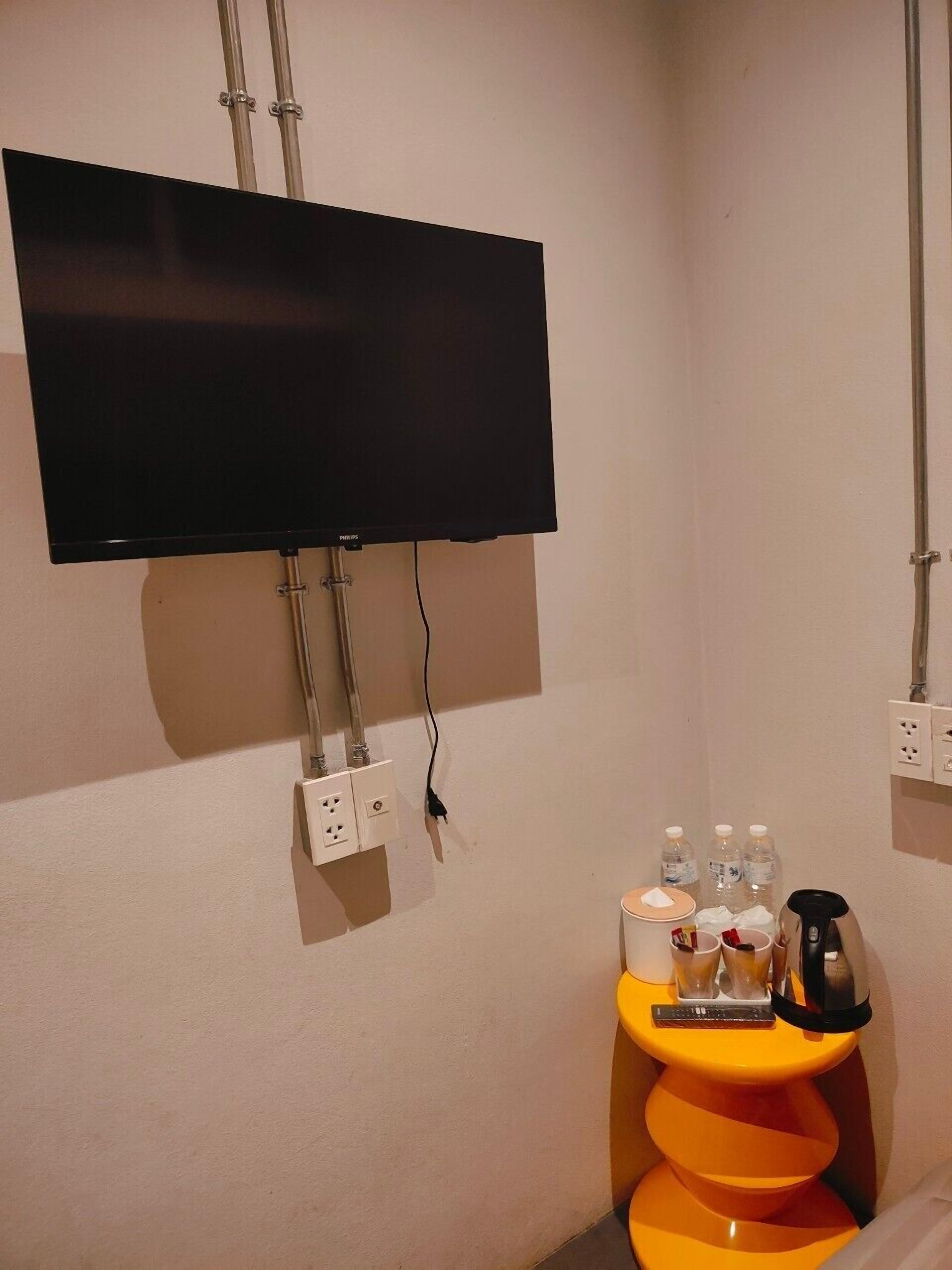 Ein Zimmer mit einem an der Wand montierten Flachbildfernseher, Steckdosen, einem kleinen orangefarbenen Tisch mit Fernbedienung, Wasserflaschen und einer Kaffeemaschine.