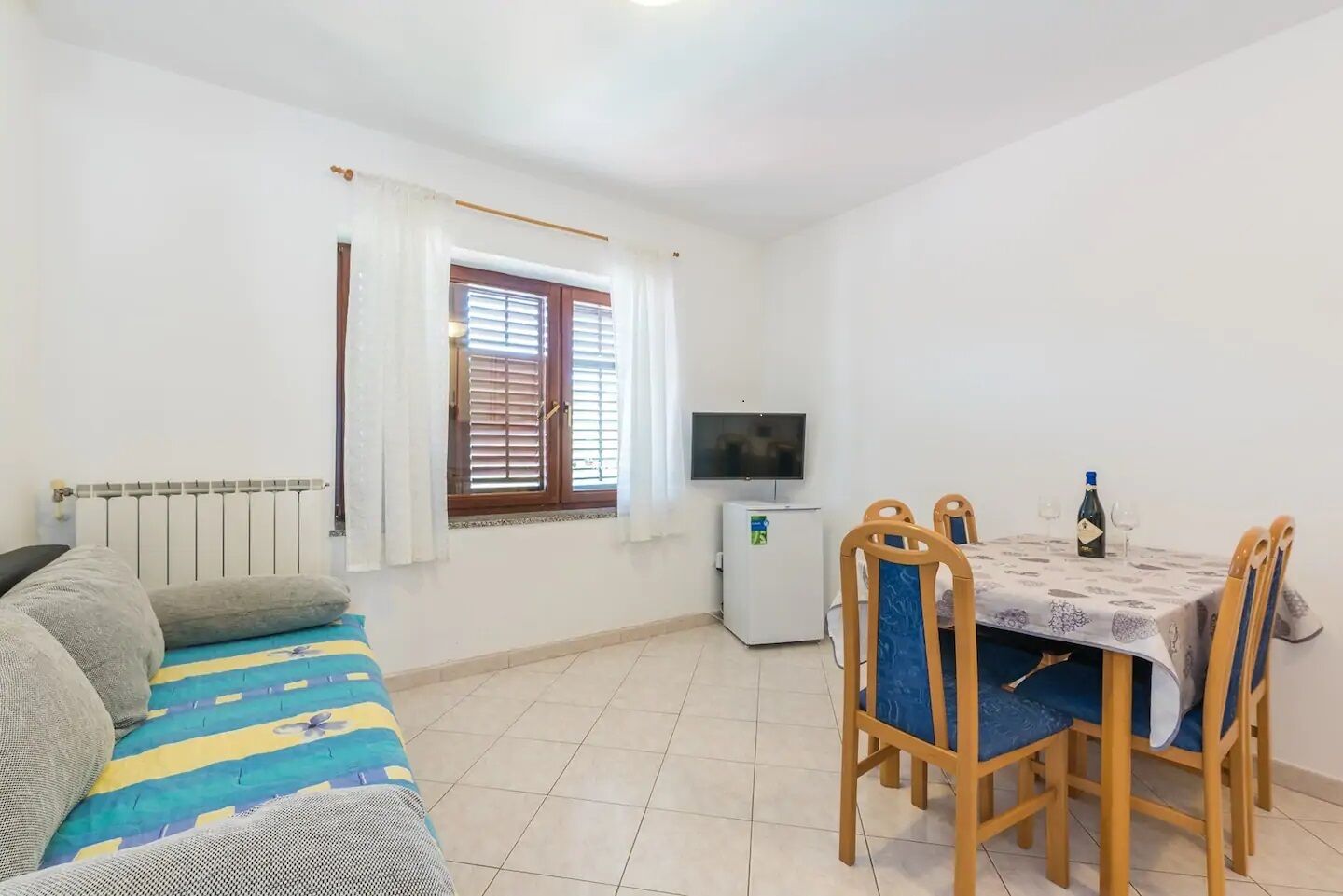 Um quarto pequeno e bem iluminado com um sofá, uma mesa de jantar com cadeiras, uma televisão e um frigorífico.