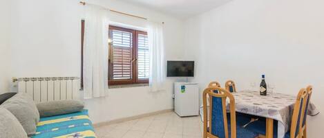 Um quarto pequeno e bem iluminado com um sofá, uma mesa de jantar com cadeiras, uma televisão e um frigorífico.