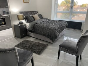 Ein modernes Schlafzimmer mit einem großen Bett, einem Nachttisch mit Lampe, einem Sessel und Blick ins Freie durch ein Fenster.