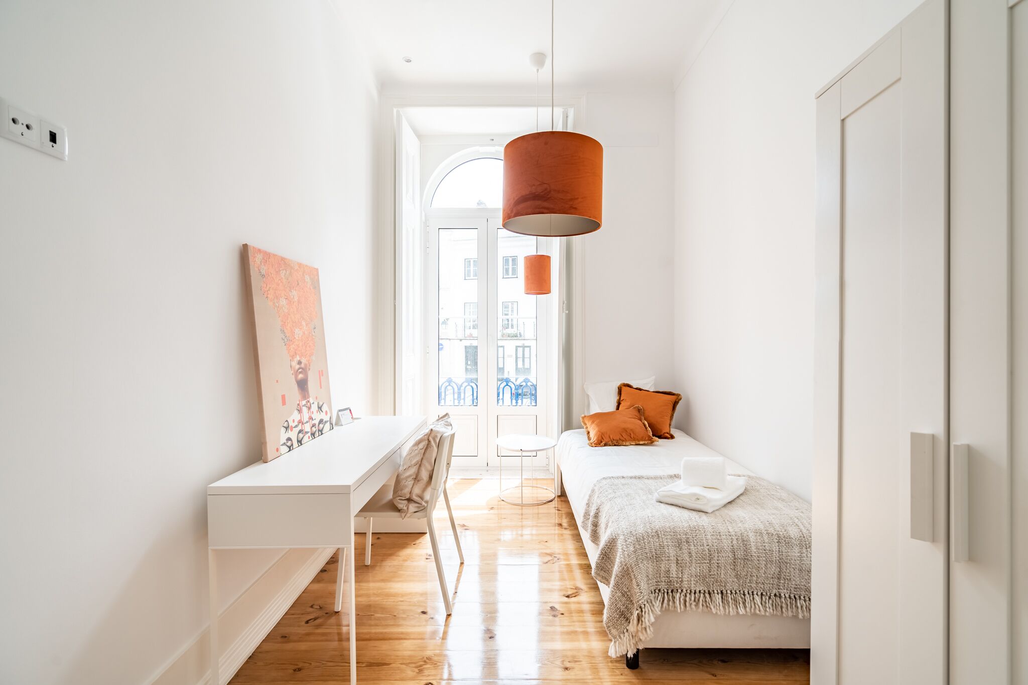 A bright, minimalist bedroom with a white desk, a bed with orange pillows, and a large window.