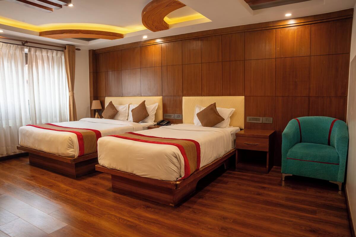 Quarto de hotel com duas camas, piso de madeira, cabeceira de madeira, mesa de cabeceira e uma poltrona azul-turquesa.
