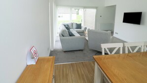 A modern living room with a grey sofa, a wooden dining table, and a TV mounted on the wall. - Harcombe House Bungalow 1 (Newton Abbot)