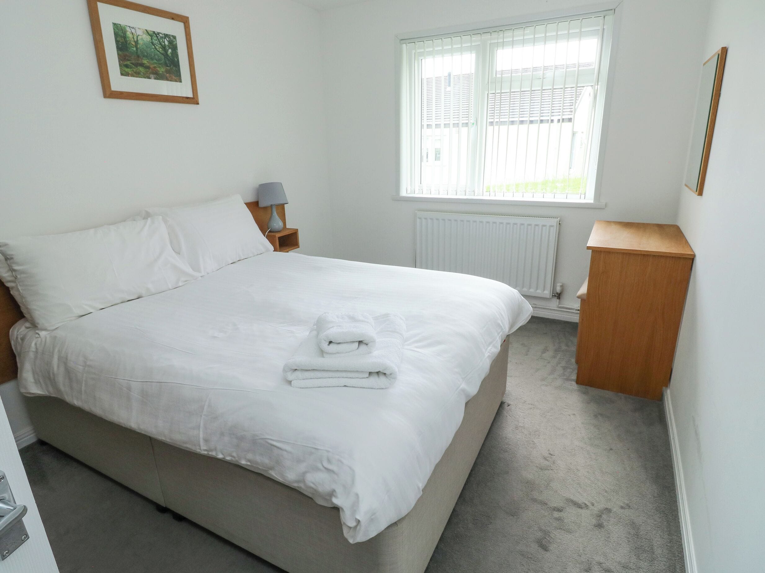 A neatly made bed with white linens and a folded towel, a bedside table with a lamp, a window with blinds, and a radiator.