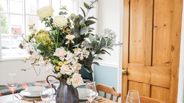 Une table à manger dressée avec de l’assiette blanche, des couverts en argent et des verres à vin, agrémentée d’un vase de fleurs.