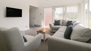 Uma sala de estar moderna com televisão de tela plana, sofás cinzas, uma mesa de centro e uma janela grande com persianas.