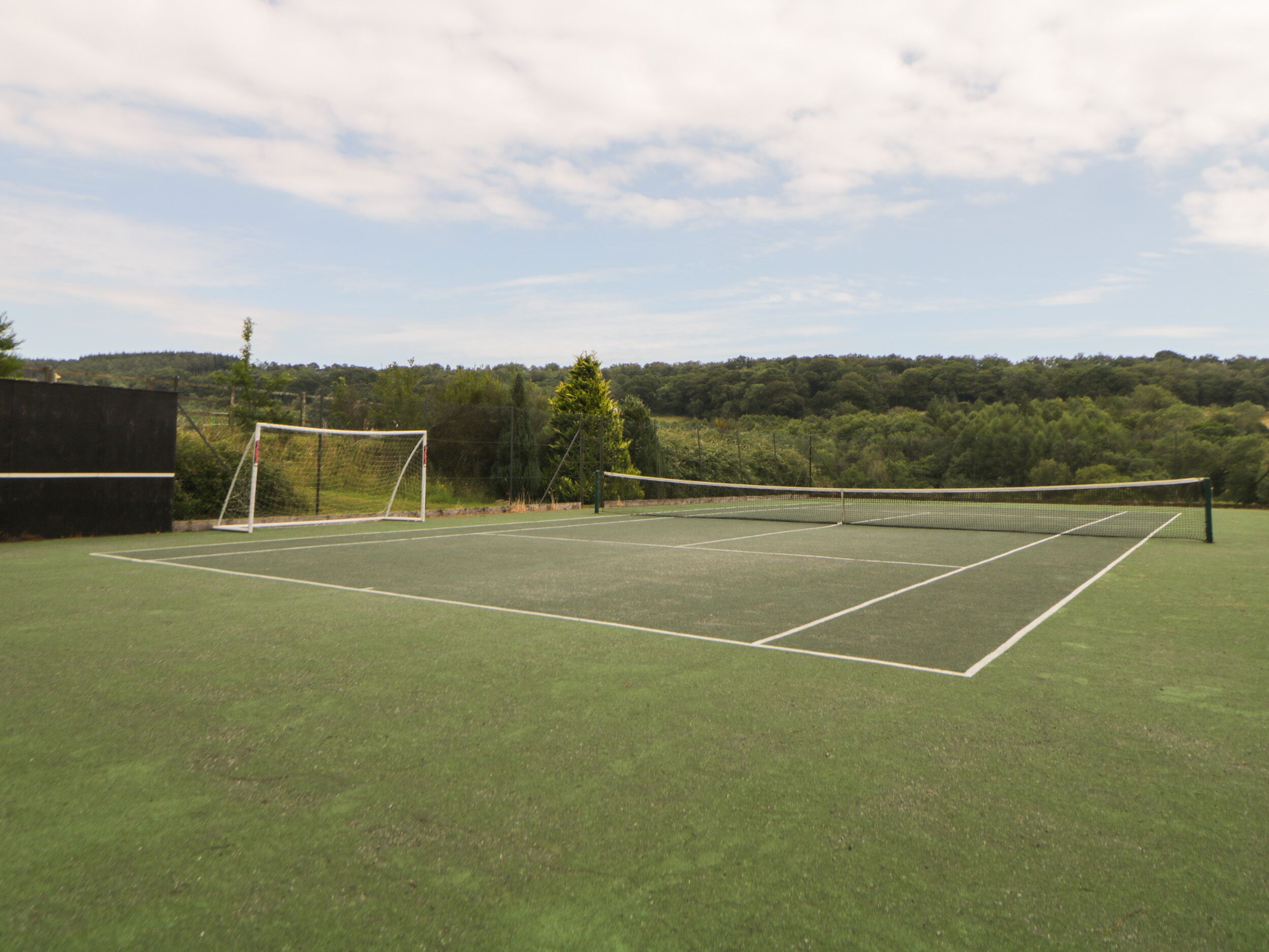 Een buitenbaan voor tennis met een net en doelpaal, omgeven door bomen en een heldere lucht.