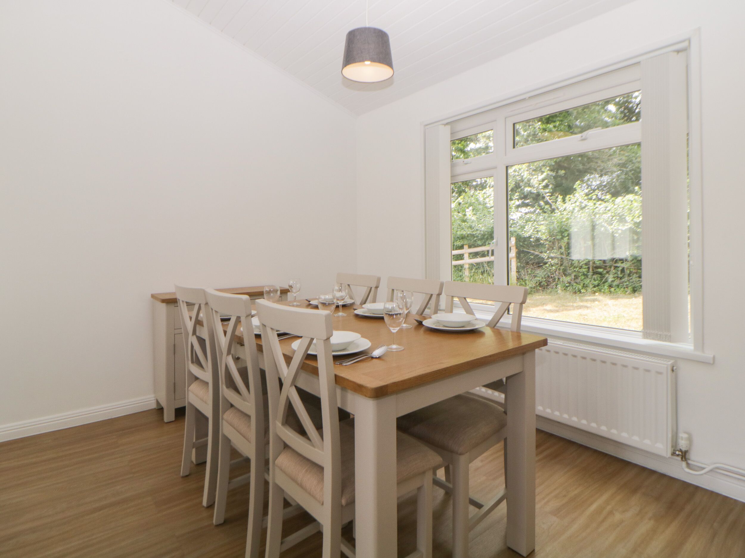 Een eetgedeelte met een houten tafel, witte stoelen en een raam met uitzicht op groen.
