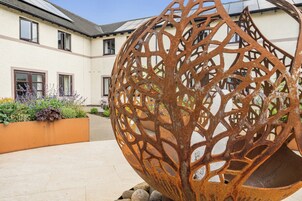 Un patio con una escultura metálica esférica, un edificio con varias ventanas y una maceta con diversas plantas.