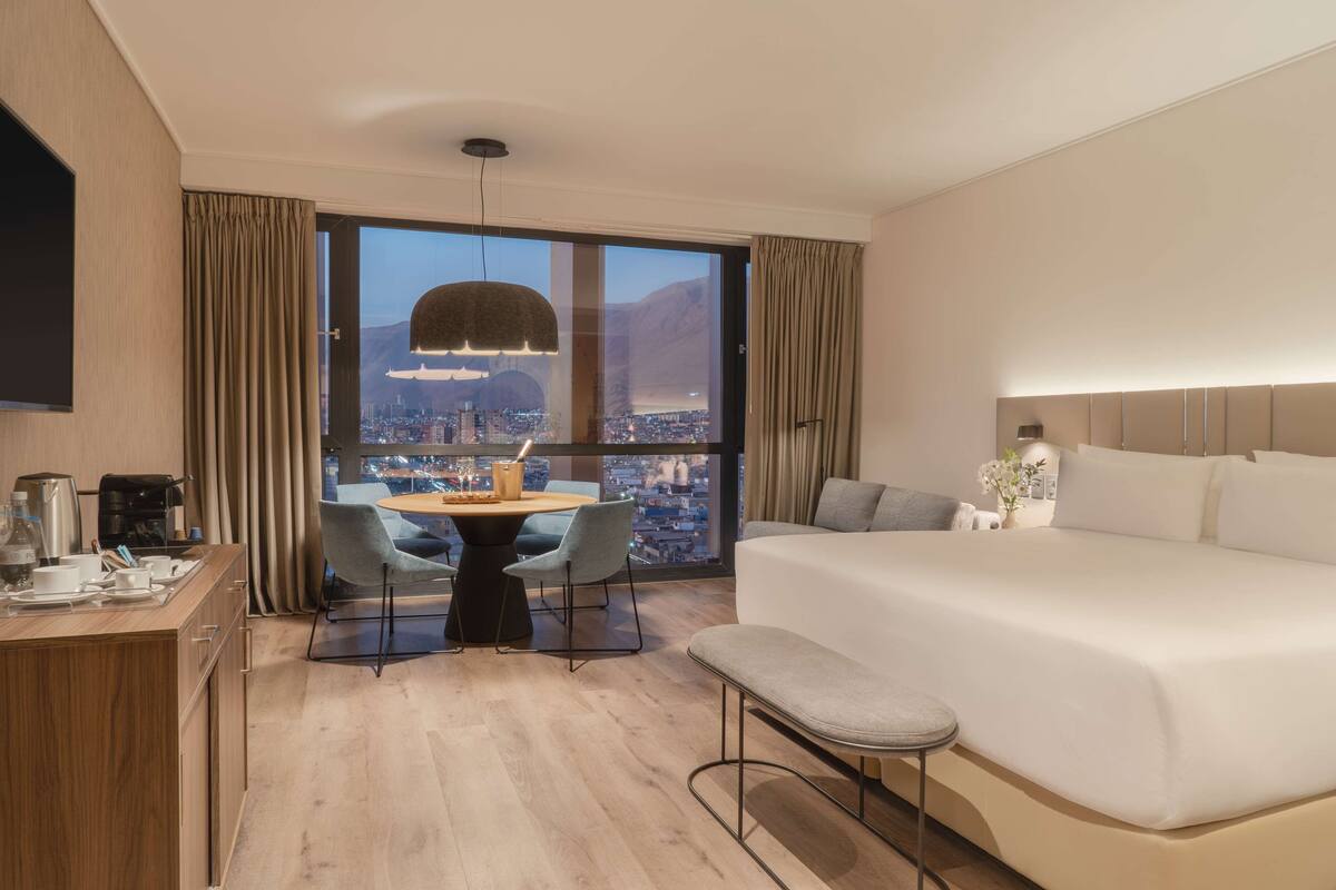 Hotelkamer met een groot bed, een bureau met koffiezetapparaat, een zithoek met stoelen en uitzicht op een stadsbeeld door grote ramen.