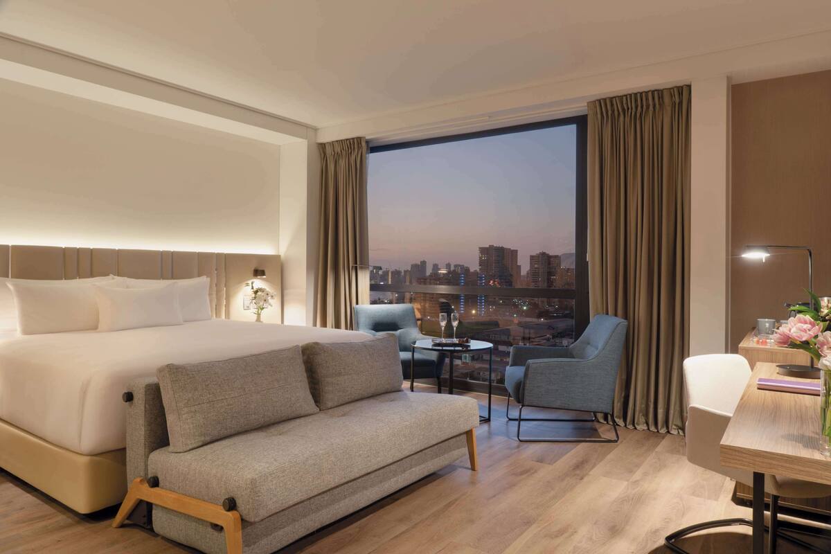 Hotelkamer met een groot bed, een bank, een bureau met lamp en uitzicht op de stad.