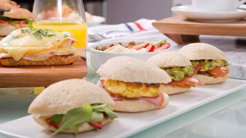 Un desayuno con sándwiches, un vaso de jugo de naranja y una taza blanca sobre una mesa.