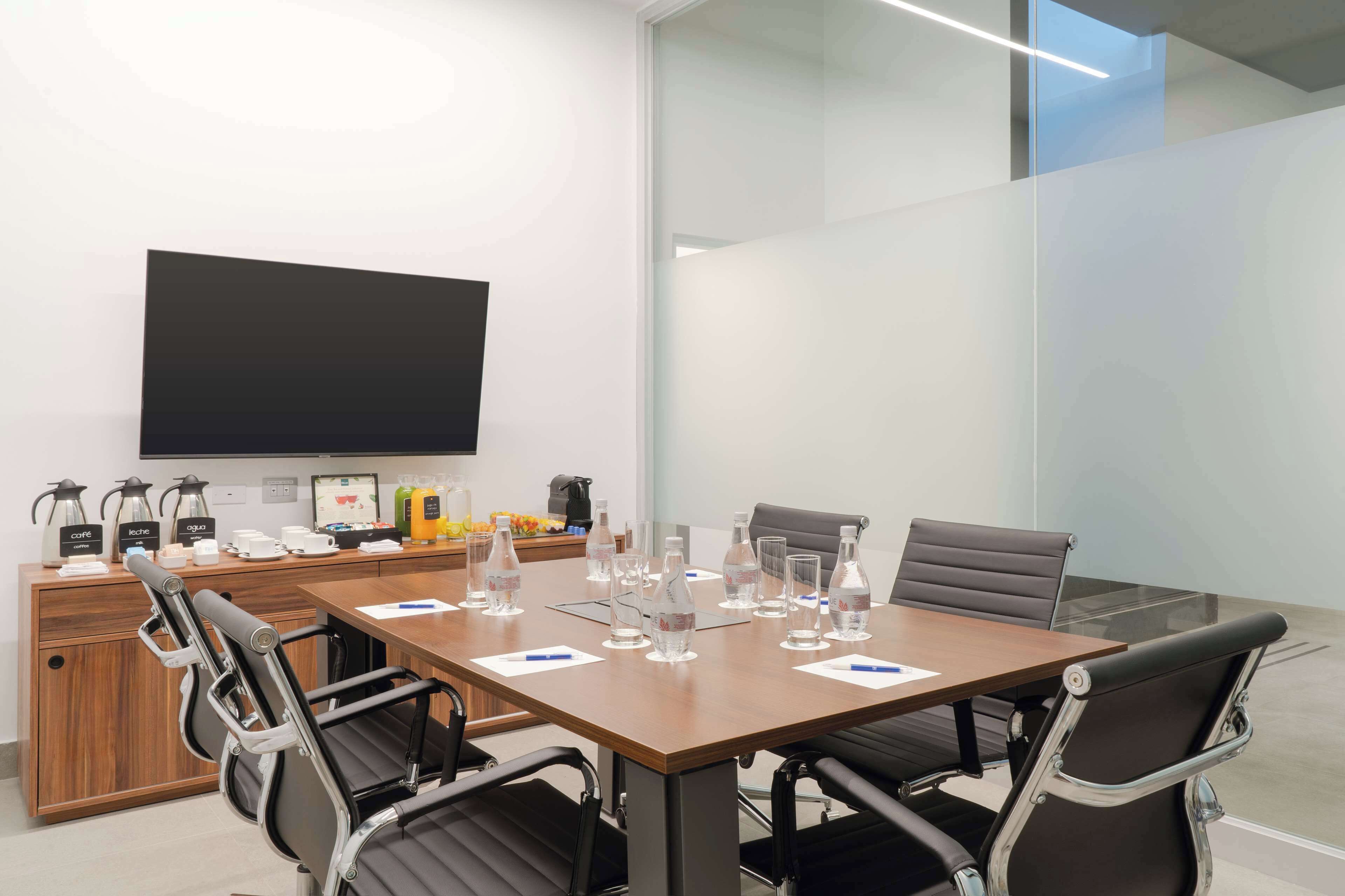 A conference room with a large table, chairs, a TV, and various amenities like coffee makers and water bottles.