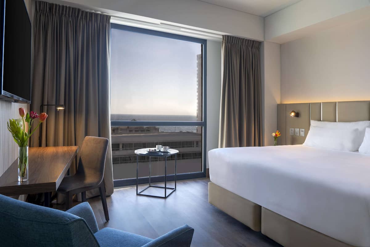 Een hotelkamer met een groot bed, een kleine tafel met een bloevas, een stoel en uitzicht op de stad door een groot raam.