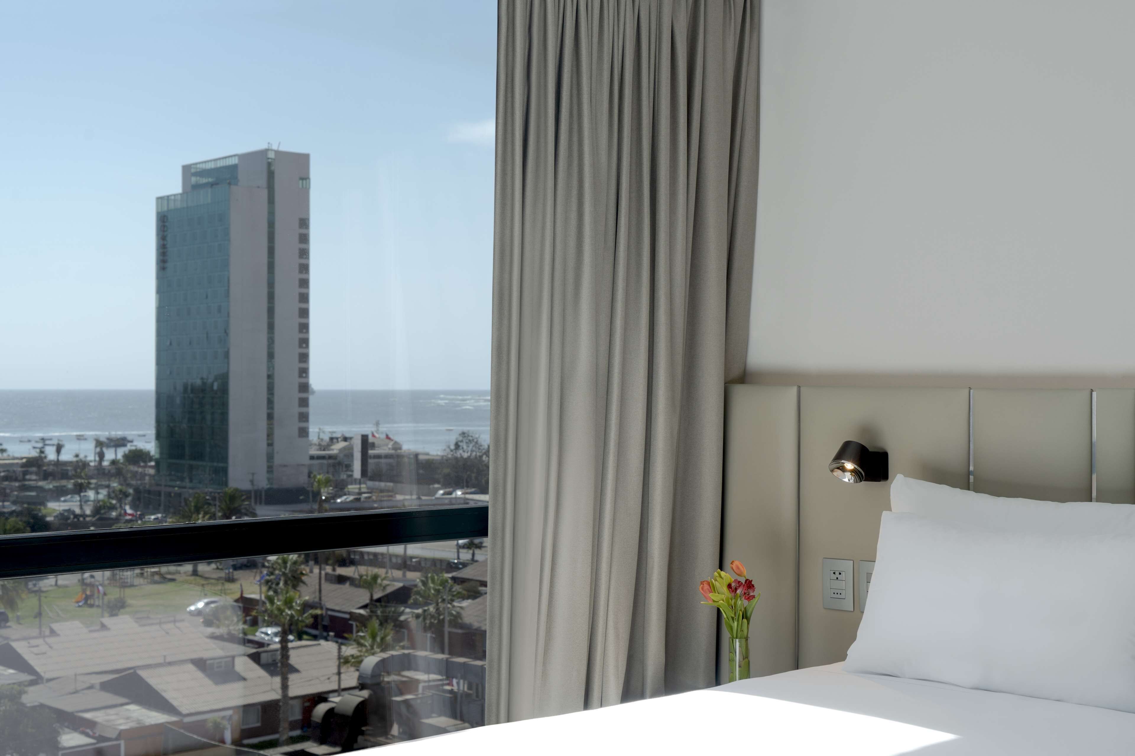 Una habitación de hotel con una cama, una mesita de noche, un jarrón con flores, una lámpara de pared y vistas a un edificio alto y al océano.
