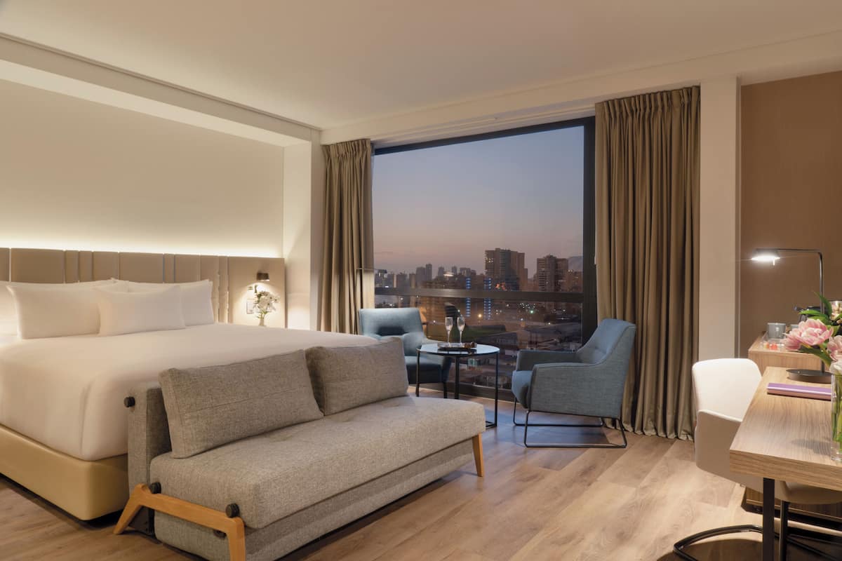 Een hotelkamer met een bed, een slaapbank, een bureau met lamp, een stoel en een groot raam met uitzicht op de stad.