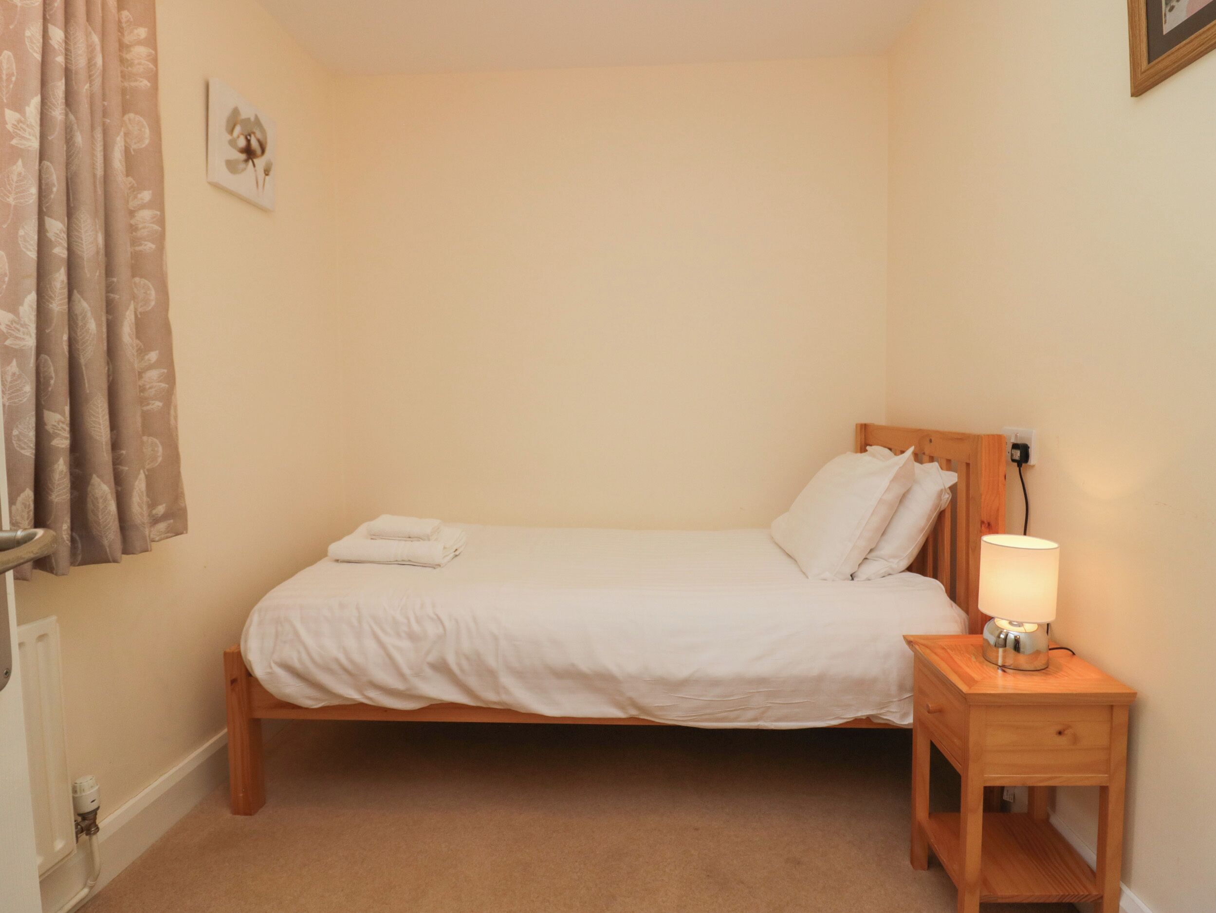 A neatly made bed with white linens and pillows, a bedside lamp, and a small wooden nightstand.