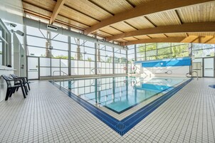 Una piscina interior con suelo de baldosas, vigas de madera en el techo y grandes ventanales.