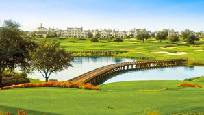 Ein Golfplatz mit einer Brücke über einem See, umgeben von üppiger Vegetation und Wohnhäusern im Hintergrund.