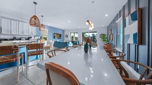 Eine moderne Küche mit weißen Schränken, ein Essbereich mit einem Glastisch und Korbstühlen sowie ein Wohnzimmer mit einem blauen Sofa und einem großen Fenster.