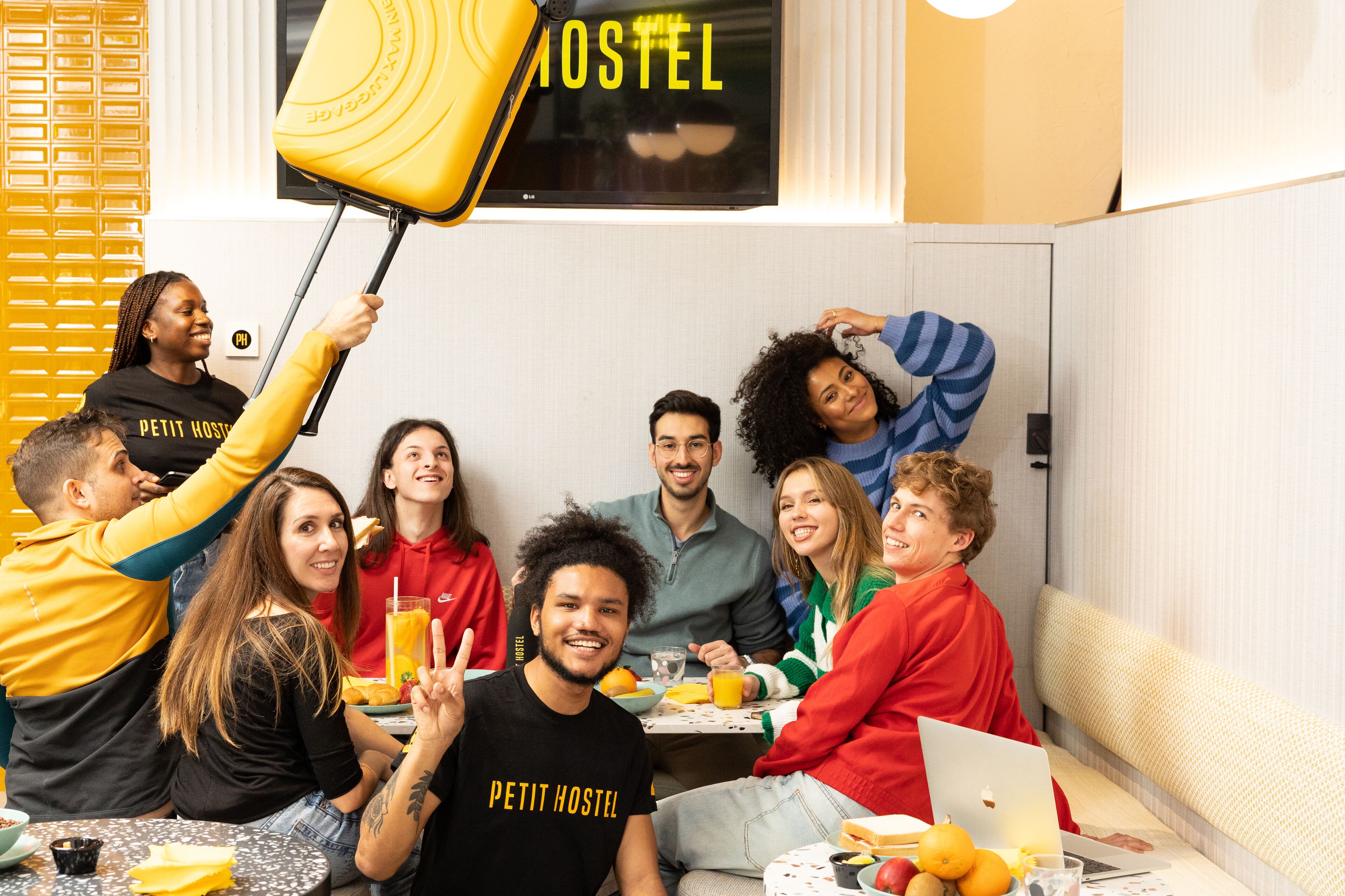 A group of people in a room with a bed, a table with food, and a TV screen displaying "HOSTEL".