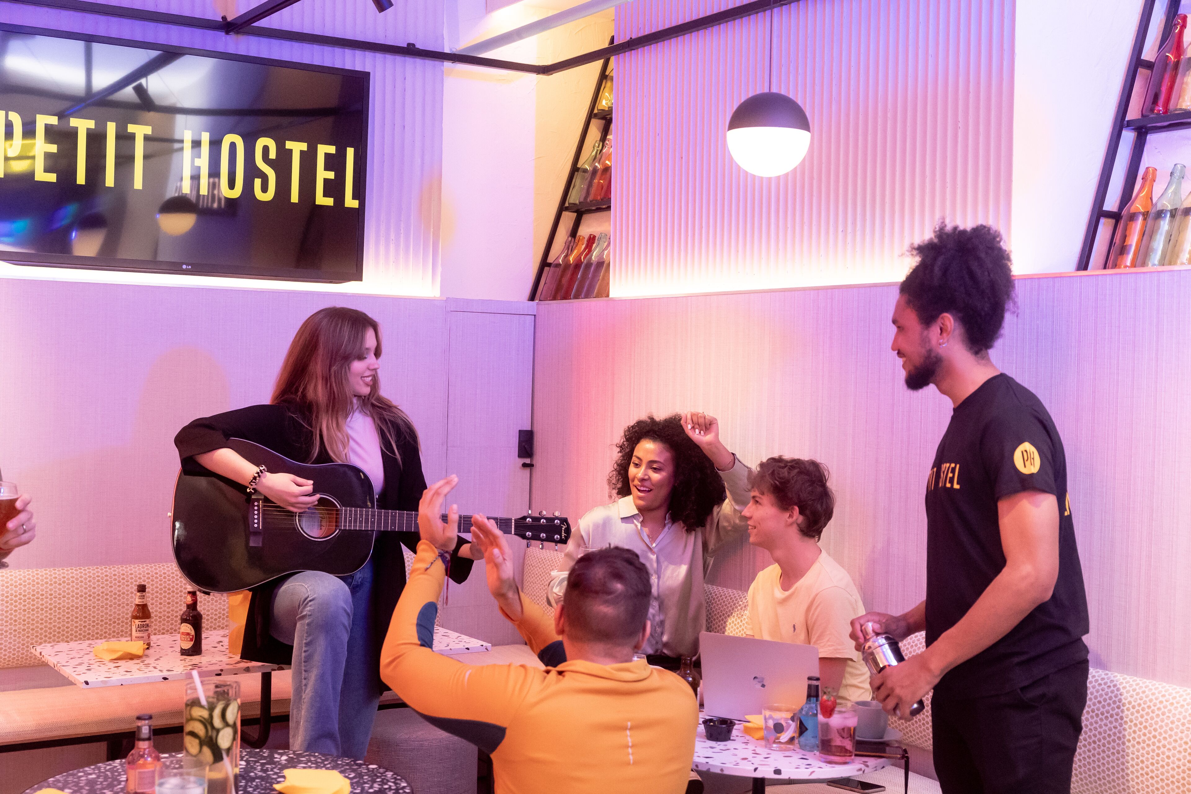 A room with a sign reading "PETIT HOSTEL," a guitar player, and people sitting and standing around a table with drinks and snacks.