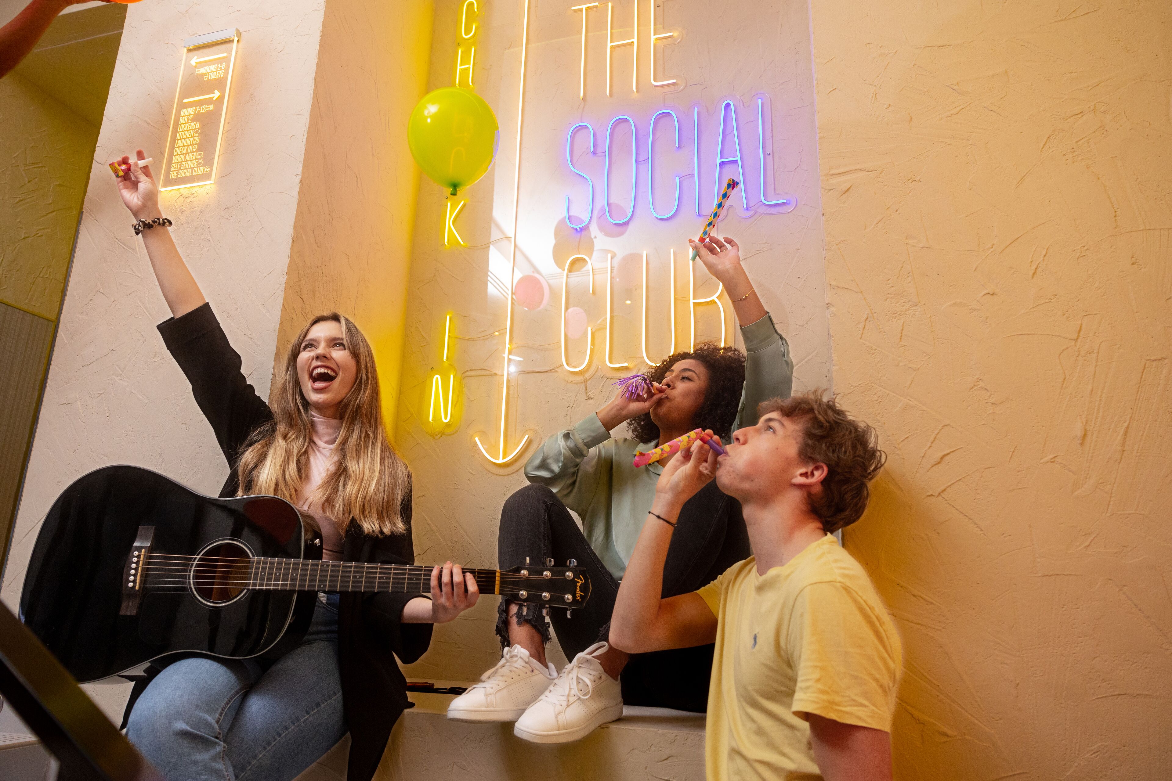 Three people sitting against a wall with a neon sign that reads "The Social Club."