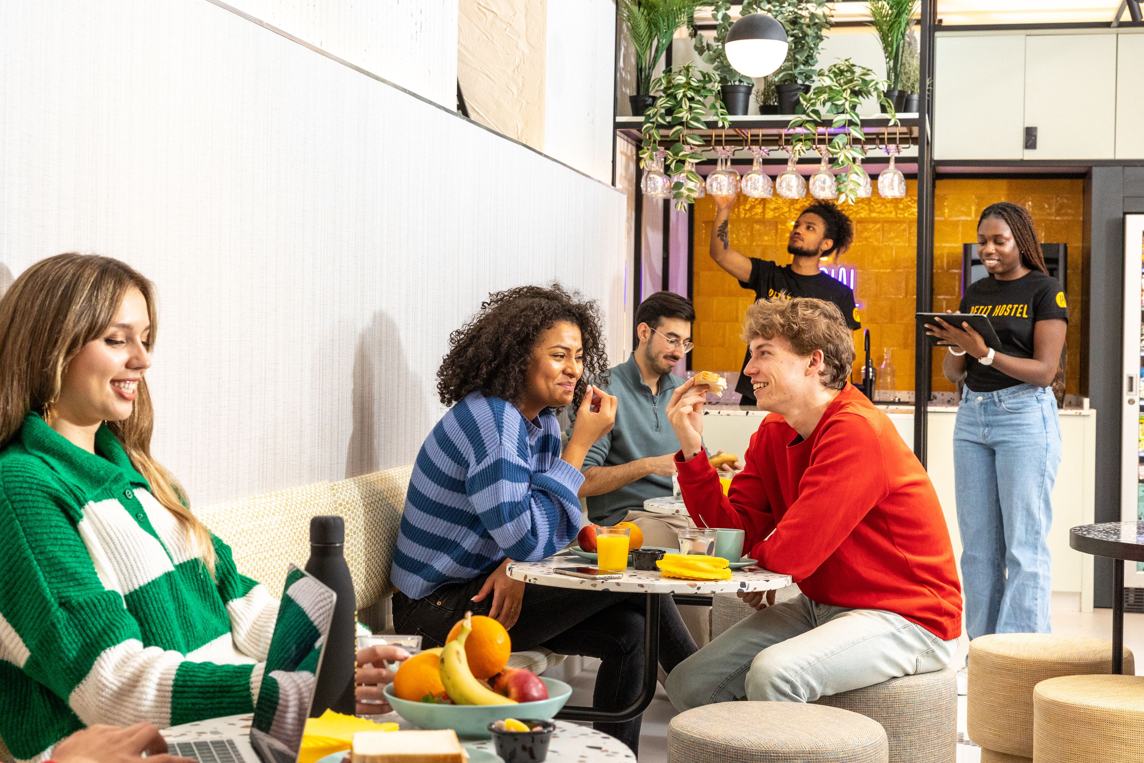 Un groupe de personnes est assis autour d’une table, devant des fruits et des boissons, dans une pièce au design moderne, agrémentée de plantes.