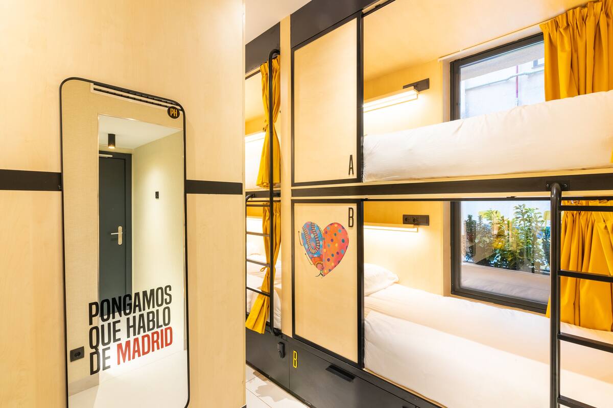 A train compartment with bunk beds, a mirror with a reflection of a door, and a window with curtains.